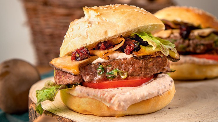 Burger mit Sesambrötchen, Salat, Tomate, Pilzen, Soße und Patty auf einer Holzplatte, im Hintergrund ein Korb mit Holz.