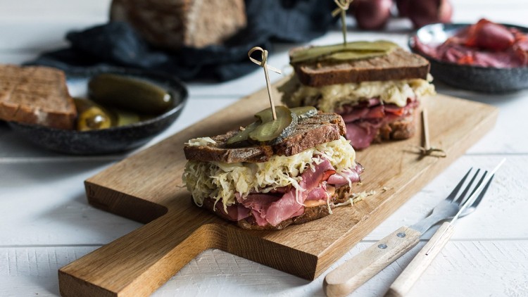 Zwei belegte Sandwiches mit Sauerkraut, Fleisch und Gewürzgurken auf einem Holzbrett, daneben Besteck und Beilagen.