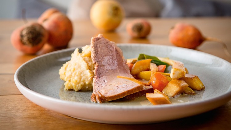 Ein Teller mit Bratenstück, Kartoffelpüree, buntem Gemüse und Soße, im Hintergrund unscharf Quitten auf einem Holztisch.