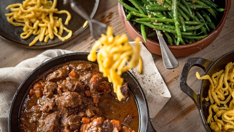Eintopf mit Fleisch und Karotten in einer Pfanne, daneben grüne Bohnen, Spätzle auf einem Teller und eine kleine Pfanne.