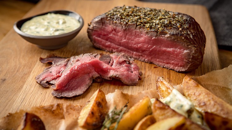 Medium gebratenes Rinderfilet mit Kräuterkruste, aufgeschnitten, daneben Kartoffelspalten und Dip auf einem Holzbrett.