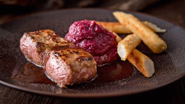 Ein gegrilltes Stück Fleisch mit rotem Püree und knusprigen Stangen auf einem dunklen Teller.