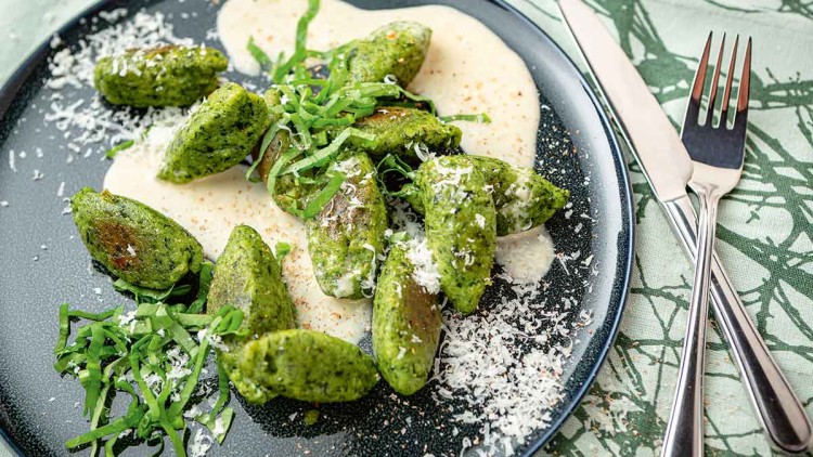 Grüne Spinatnocken auf heller Soße liegen auf einem dunklen Teller, garniert mit geriebenem Käse und frischem Basilikum.