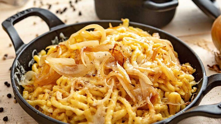 Pfanne mit Spätzle, geschmolzenem Käse und angerösteten Zwiebeln, dahinter grüne Beilage in einer kleinen schwarzen Schale.