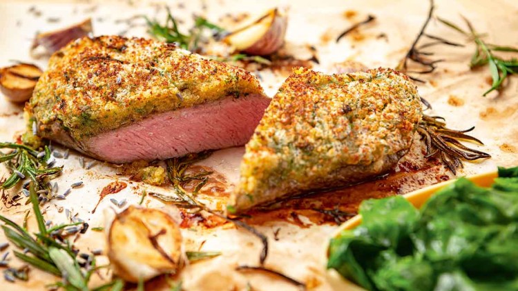 Ein Stück rosa gebratenes Fleisch mit Kräuterkruste liegt auf Backpapier, dekoriert mit Rosmarin und Knoblauch, daneben Blattspinat.