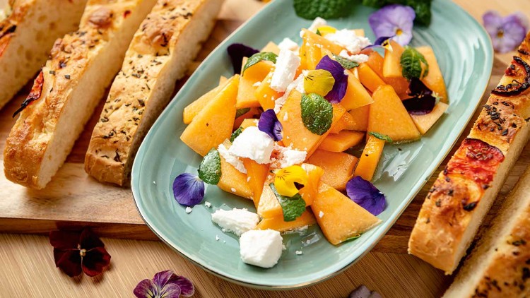 Obstsalat mit Melonenstücken, Minzblättern, Blüten und Fetakäse auf einem Teller, daneben Kräuter-Focaccia-Scheiben.