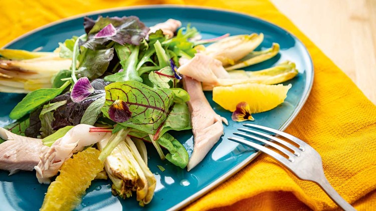 Grüner Blattsalat mit Chicorée, Streifen von geräuchertem Fisch, Orangenspalten und essbaren Blüten auf einem blauen Teller.