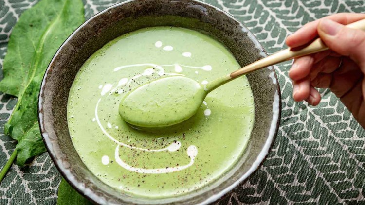 Grüne Suppe mit Sahnetupfen und Pfeffer in einer Schale, ein Löffel wird von einer Hand hineingetaucht, Blatt im Hintergrund.