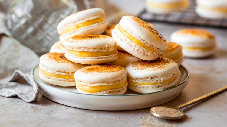 Weiße Macarons mit gelber Cremefüllung auf einem Teller, daneben Trockenblumen in einer Vase und ein goldener Teelöffel.