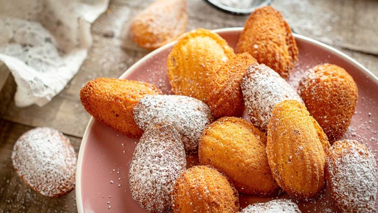 Mehrere goldbraune Madeleines liegen mit Puderzucker bestäubt auf einem rosa Teller, daneben Holz und ein weißes Tuch.