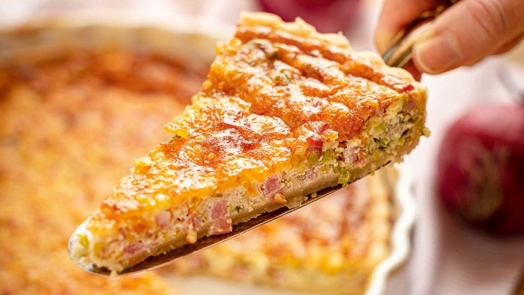 Eine Hand hebt ein Stück Quiche mit Käse und Schinken aus einer weißen runden Auflaufform, im Hintergrund rote Zwiebeln.