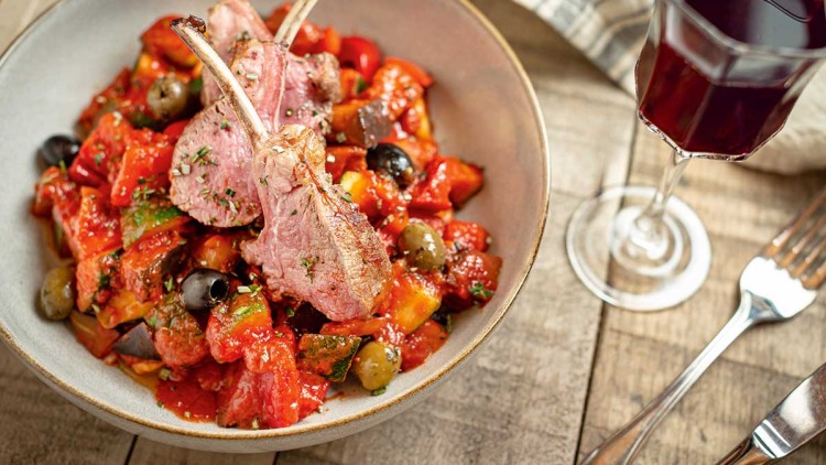Grauer Teller mit zwei Lammkoteletts auf Ratatouille aus buntem Gemüse, Oliven und Tomatensoße, daneben ein Glas Rotwein.