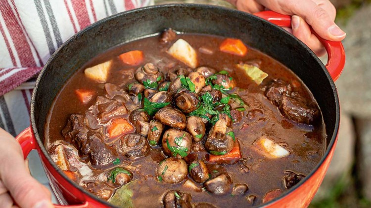 Rote Gusseisenpfanne mit Eintopf aus Pilzen, Karotten, Fleisch und Petersilie wird von zwei Händen gehalten.