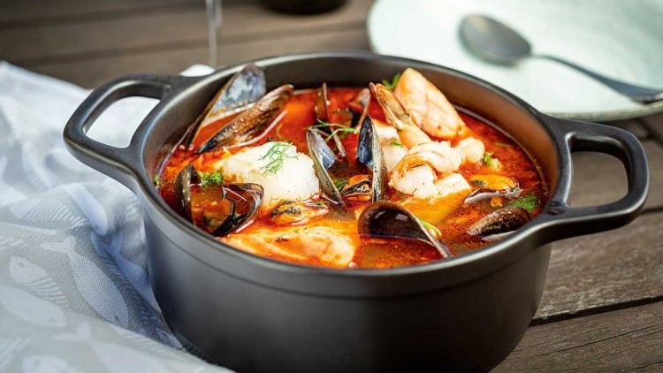 Schwarzer Topf mit roter Fischsuppe, Garnelen, Miesmuscheln und Kräutern auf Holztisch, daneben ein Glas Weißwein und ein Teller.