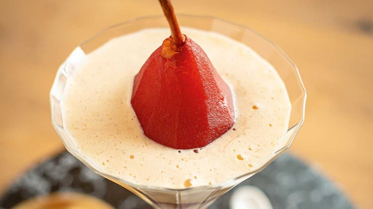Eine rote gedünstete Birne steht in einer cremefarbenen Dessertcreme in einem Glas, daneben Kekse auf einem goldenen Teller.