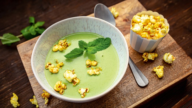 Grüne Cremesuppe mit Minzblättern und gelbem Popcorn in einer Schale, daneben ein Löffel und ein Becher mit Popcorn.