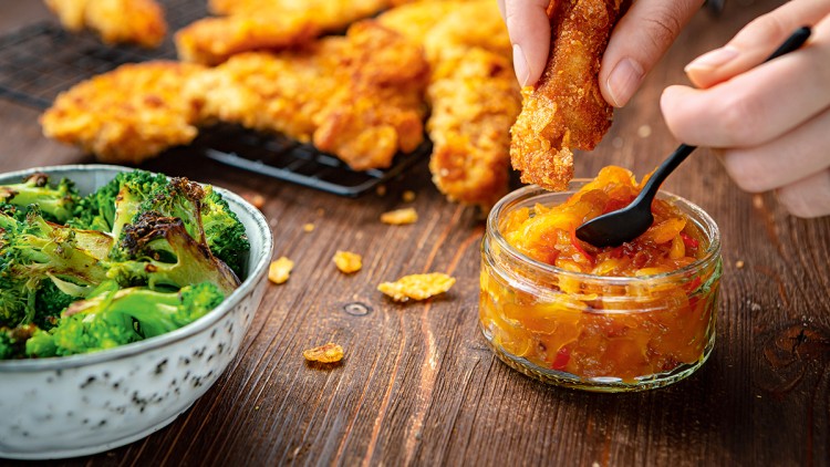 Hand taucht ein knuspriges Hähnchenstück in ein Glas mit orangem Dip, daneben Schale mit geröstetem Brokkoli und Hähnchen auf Gitter.
