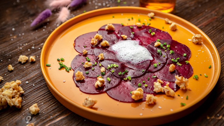 Rote-Bete-Carpaccio mit Walnussstücken, Schnittlauch, Schaum und Salzflocken auf einem orangefarbenen Teller.