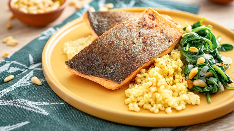 Zwei gebratene Fischfilets mit knuspriger Haut, gelbem Couscous, frischem Blattspinat und Pinienkernen auf gelbem Teller.