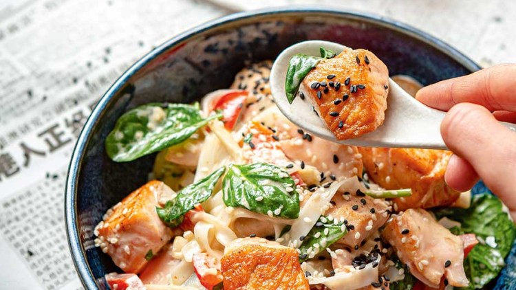 Bunte Nudelschale mit Lachs, Spinat, Paprika, Sesam und Cremesauce auf Zeitung, daneben rote Chilischoten in Holzschale.