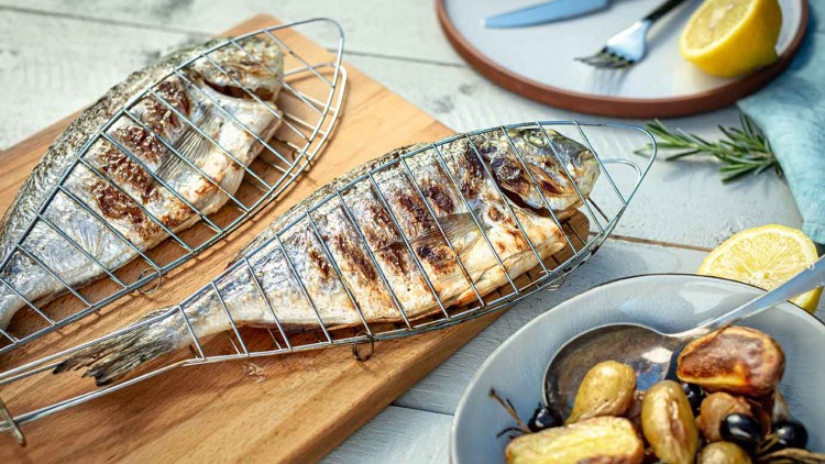 Zwei gegrillte Fische in Metallhaltern auf Holzbrett, daneben ein Teller mit Kartoffeln und Oliven sowie angeschnittene Zitronen.