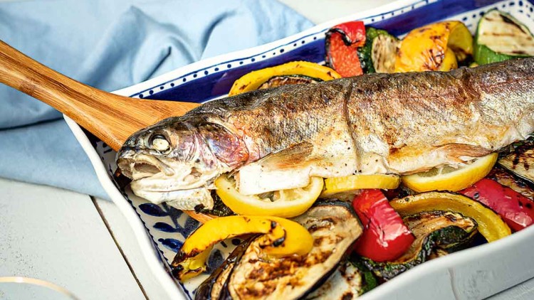 Ganze gebratene Forelle auf gegrilltem Gemüse wie Paprika, Zucchini und Aubergine mit Zitronenscheiben angerichtet.