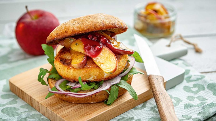 Ein Burger mit paniertem Käse, Apfelscheiben, Rucola, roten Zwiebeln und Sauce auf einem Holzbrett, ein Messer liegt daneben.