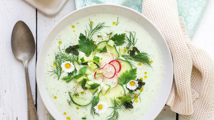 Ein Teller mit grüner Suppe, garniert mit Gurken, Radieschen, Kräutern und Gänseblümchen, daneben Brötchen und Gemüse.