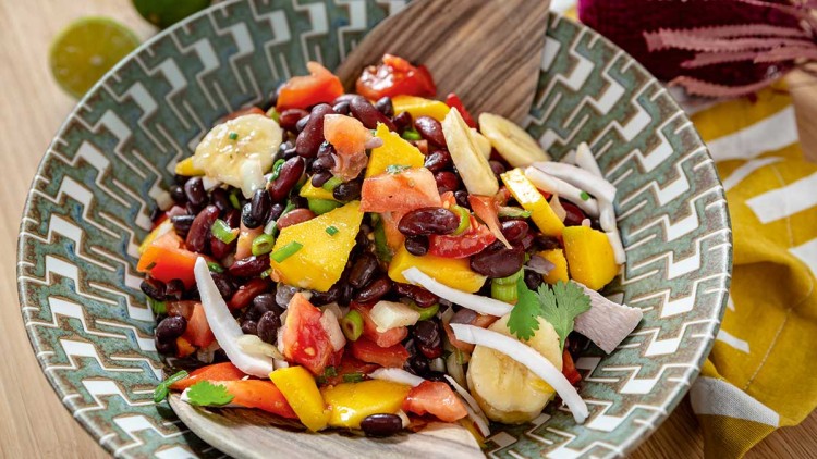 Bunte Obst- und Bohnensalat-Mischung mit Mango, Banane, Tomaten und Kräutern in einer grün gemusterten Schale mit Holzlöffel.