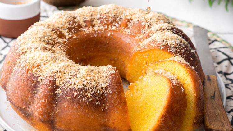 Ein goldbrauner Kuchen mit einer fehlenden Scheibe wird mit Flüssigkeit aus einer kleinen hellen Kanne begossen.