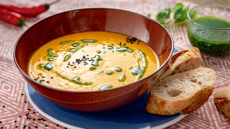 Rote Schüssel mit gelber Suppe, garniert mit grüner Soße und schwarzen Sesamsamen, daneben Baguettescheiben auf Teller.
