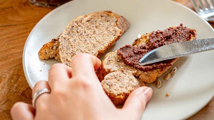 Eine Hand streicht Schokoladencreme mit einem Messer auf ein Scheibe Körnerbrot auf einem Teller, daneben Kaffee und Waffeln.