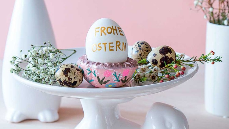 Weiße Etagere in Hasenform mit verzierten Ostereiern, Wachteleiern, kleinen Blumen und der Aufschrift „Frohe Ostern“.