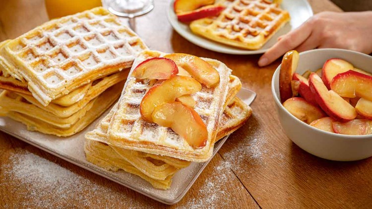 Gestapelte Waffeln mit Puderzucker und Apfelscheiben auf einem Tisch, daneben ein Teller, ein Glas Saft und eine Schüssel mit Obst.