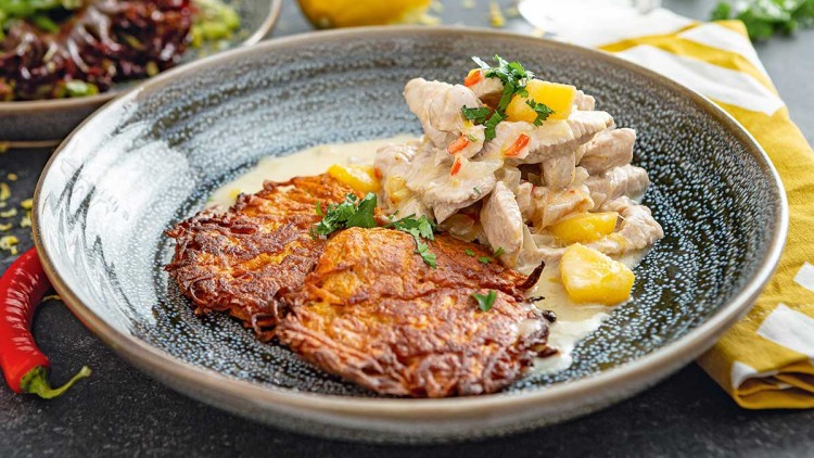 Ein Teller mit zwei goldbraunen Reibekuchen, Hähnchestreifen in Sahnesoße, Ananasstücken und frischer Petersilie.
