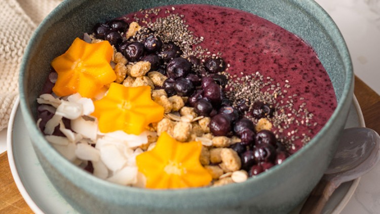 Eine Schale mit roter Smoothie-Basis, dekoriert mit Chiasamen, Blaubeeren, Granola, Kokosflocken und gelben Sternfruchtscheiben.