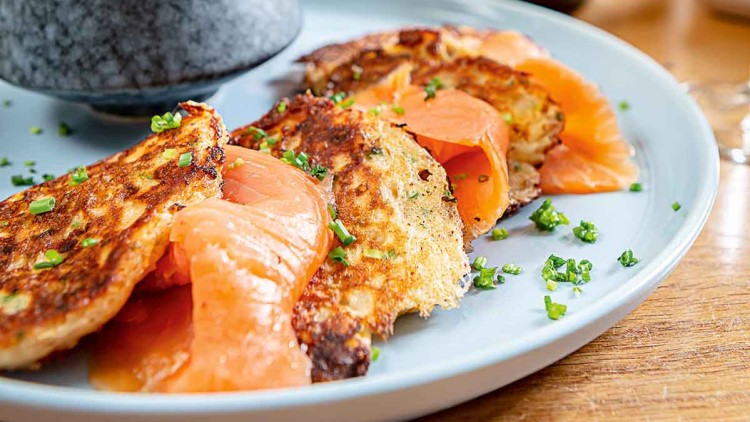 Zwei Kartoffelpuffer mit Räucherlachs, garniert mit Schnittlauch, auf einem blauen Teller neben einer Schale Avocadodip.