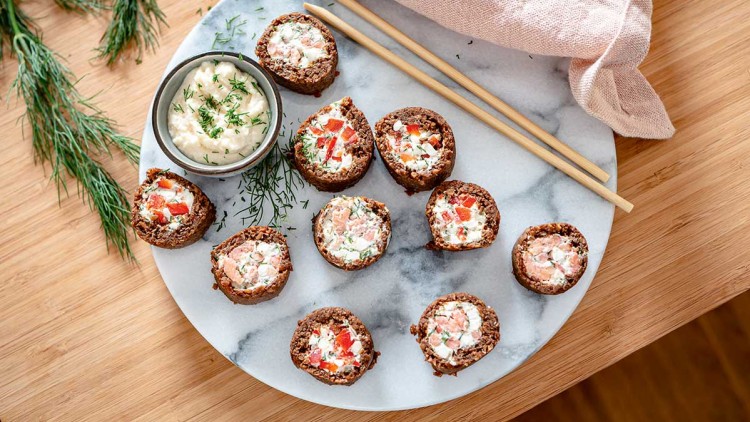 Röllchen mit Frischkäse, Lachs und Paprika auf einer weißen Platte, daneben eine Schale Dip, Dill und zwei Holzstäbchen.