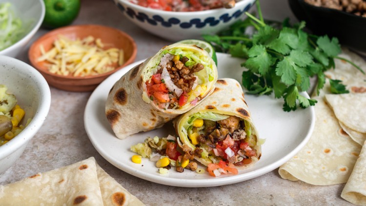 Zwei aufgeschnittene Wraps mit Gemüse, Mais und Hackfleisch auf einem Teller, umgeben von Zutaten wie Koriander und Käse.