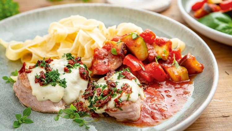 Ein Teller mit Bandnudeln, überbackenem Schweinefilet mit Kräutern und Tomaten sowie Ratatouille aus Zucchini und Paprika.