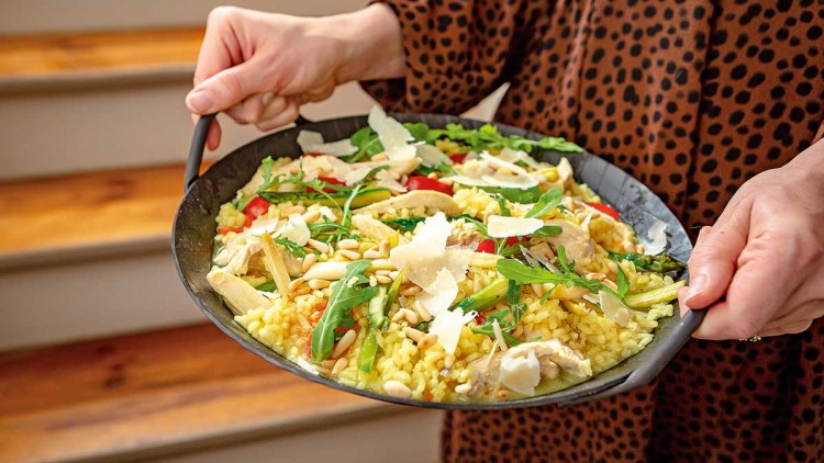 Eine Person hält eine Pfanne mit gelbem Reis, Hähnchenstreifen, Rucola, Kirschtomaten, Spargel, Pinienkernen und Parmesan.