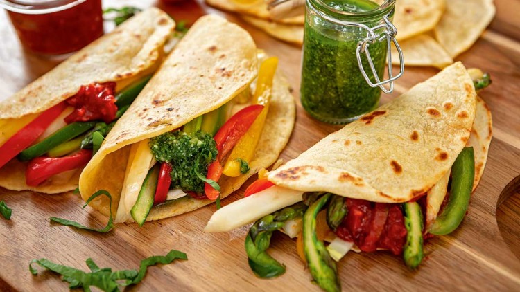 Drei mit Gemüse wie Paprika, Spargel und Sauce gefüllte Wraps liegen auf einem Holzbrett neben einem Glas mit grüner Sauce.