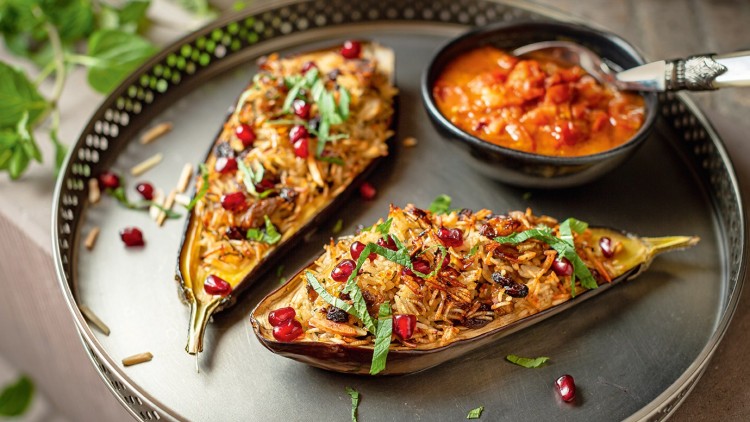 Zwei gefüllte Auberginenhälften mit Reis, Granatapfelkernen und Kräutern auf einem Tablett, daneben Schale mit Tomatensauce.