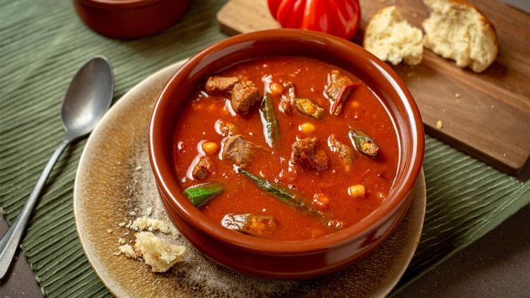 Roter Eintopf mit Fleischstücken, grünen Bohnen und Maiskörnern in einer braunen Schale, daneben Brot, Tomate und Löffel.