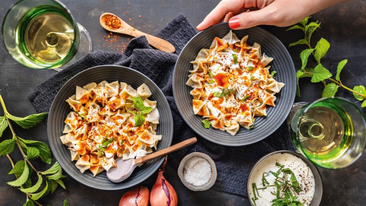 Zwei Schalen gefüllt mit gefalteten Teigtaschen, Joghurt, rotem Gewürz und Minze, daneben Schalotten, Soße und Gläser mit hellem Getränk.