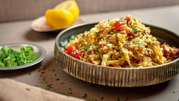 Schüssel mit Bulgurgericht, Tomaten, gehackten Kräutern und Kohl, daneben Teller mit Petersilie und halbierte Zitrone im Hintergrund.