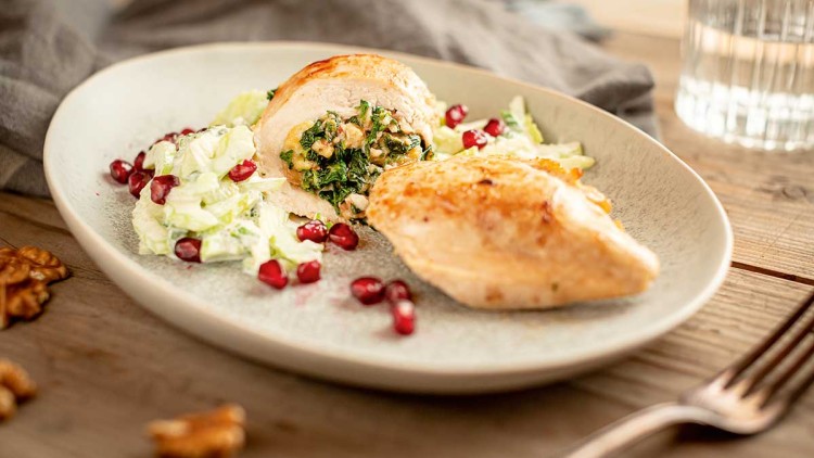 Gefüllte Hähnchenbrust mit grünem Blattgemüse und Nüssen, serviert mit Salat, Granatapfelkernen und grauer Stoffserviette.