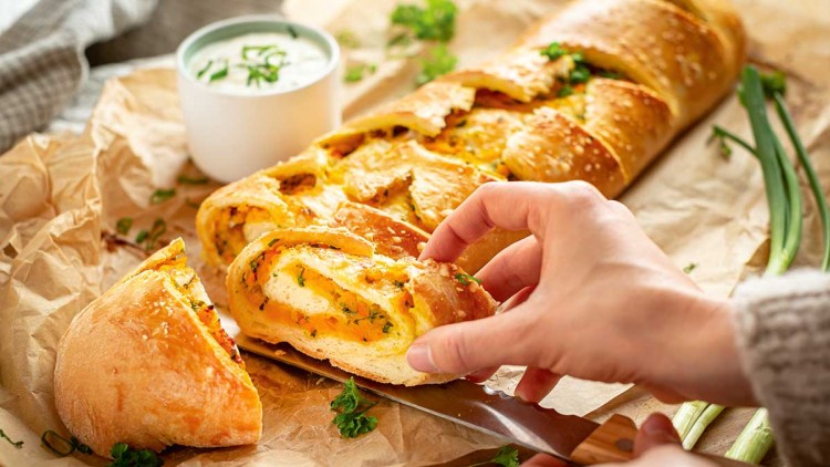 Eine Hand schneidet ein goldbraunes, gefülltes Brot mit Kräutern auf Backpapier, daneben ein Becher mit Kräuter-Dip.