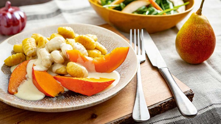 Ein Teller mit Gnocchi, Kürbisspalten und heller Soße steht neben Besteck, einer Birne und einer Schüssel Salat auf einem Tisch.
