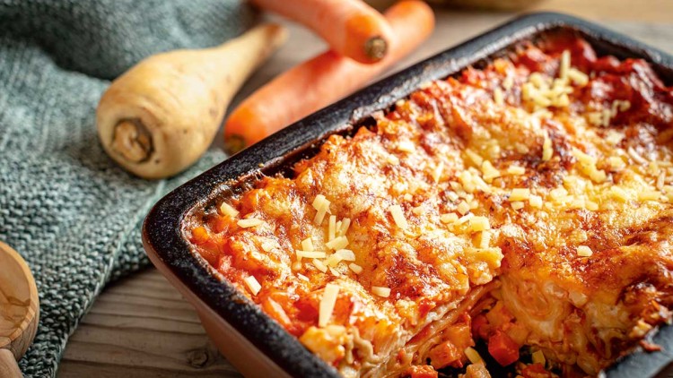 Eine Gemüse-Lasagne in einer schwarzen Auflaufform mit gebräuntem Käse, daneben Karotten, Pastinake und ein gestricktes Tuch.